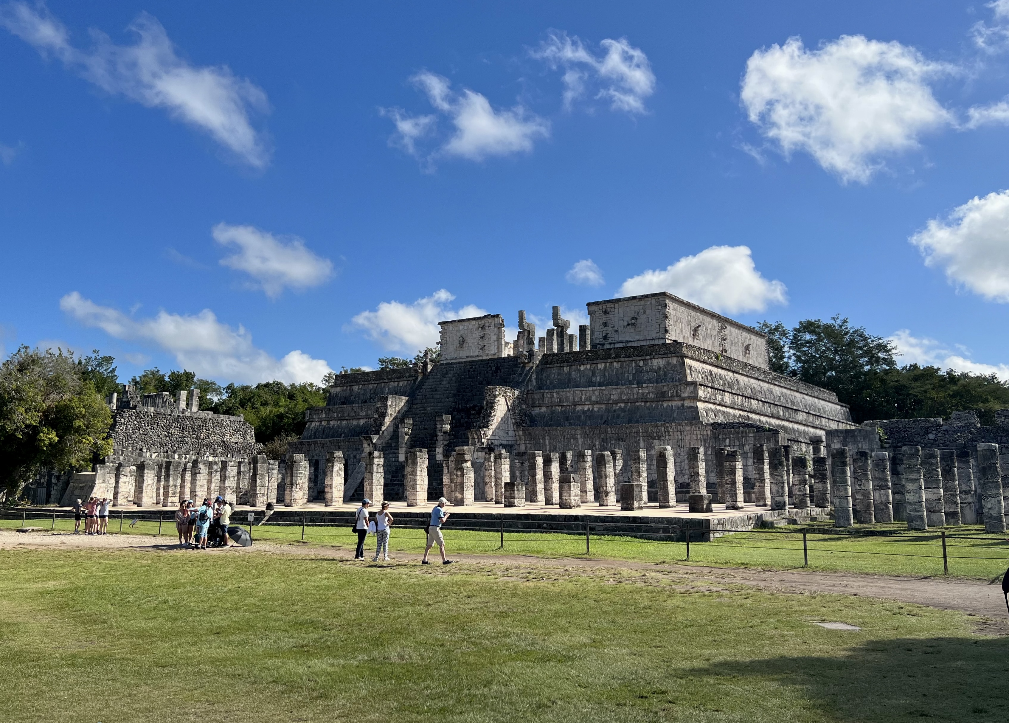 Temple of the Warriors at Chichen Itza with rows of columns, tourists exploring the site, and a bright blue sky with clouds above