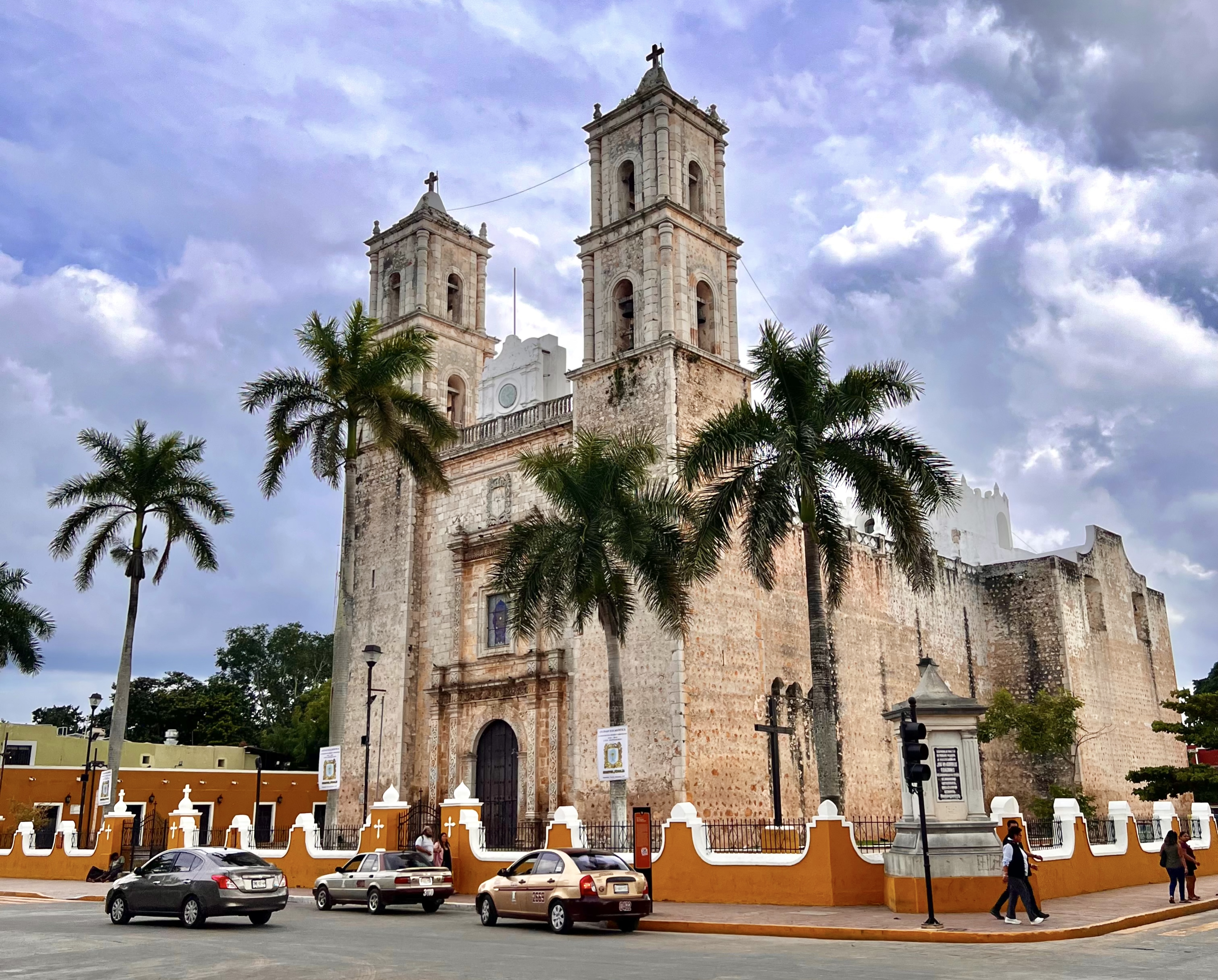 "Colonial-style church in Valladolid, Mexico, with twin bell towers, surrounded by palm trees, cars, and a cloudy sky in the background