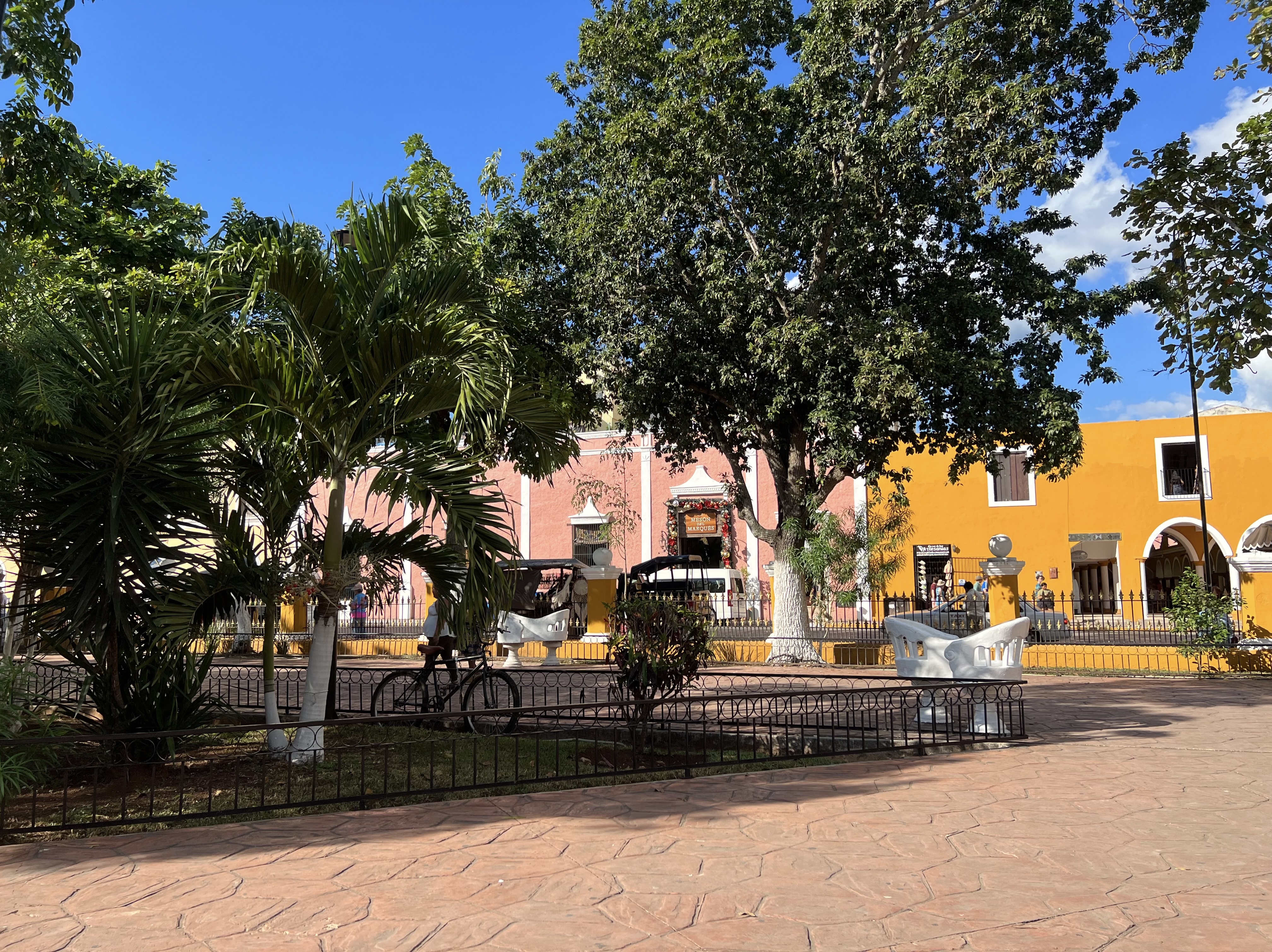 Sunny park in Valladolid, Mexico, with lush trees, benches, and a view of colorful buildings in the background, including pink and yellow facades